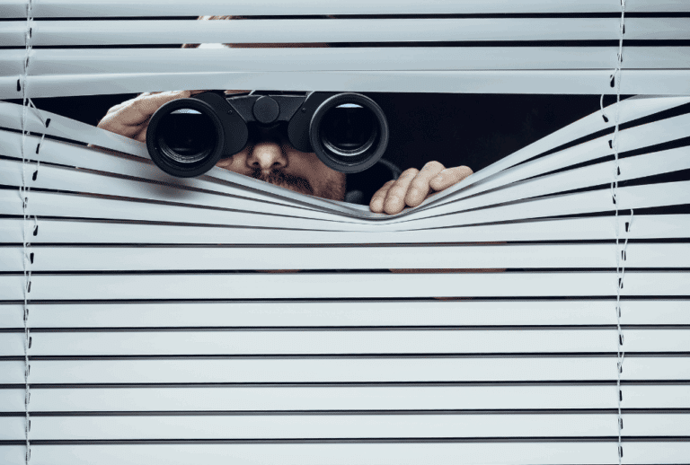 Image is of a person conspicuously peering through window blinds with binoculars, concept of DIY surveillance in Seattle