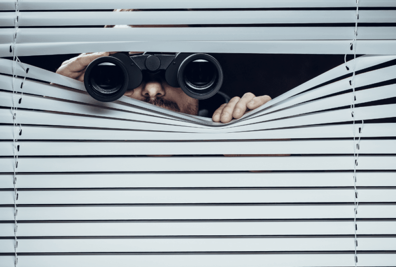 Image is of a person conspicuously peering through window blinds with binoculars, concept of DIY surveillance in Seattle
