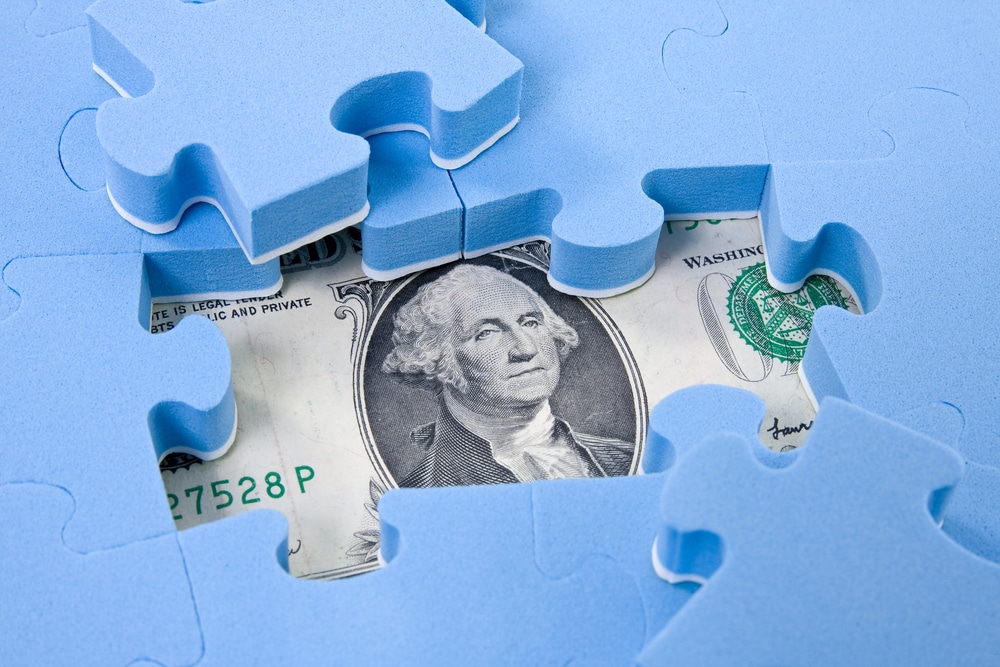 Hidden dollar bill under puzzle pieces representing uncovering hidden bank accounts.