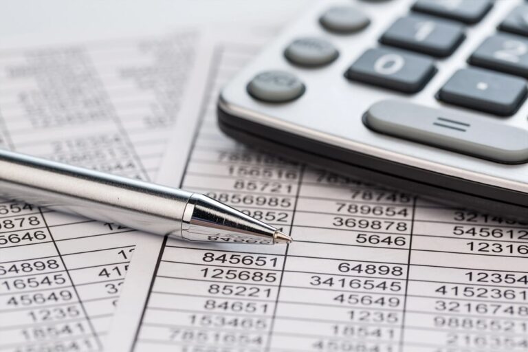 Image displays a pen, calculator, and printed number sheets on a desk, concept of how an asset search analyzes financial data to confirm assets and liabilities.