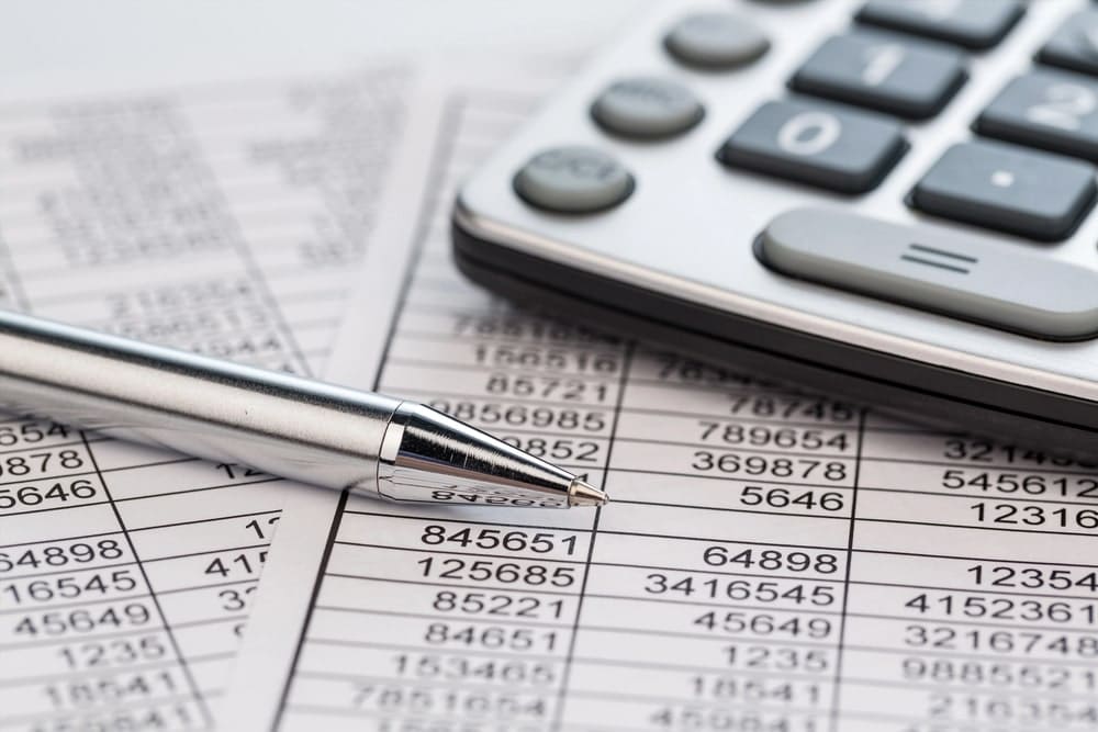 Image displays a pen, calculator, and printed number sheets on a desk, concept of how an asset search analyzes financial data to confirm assets and liabilities.