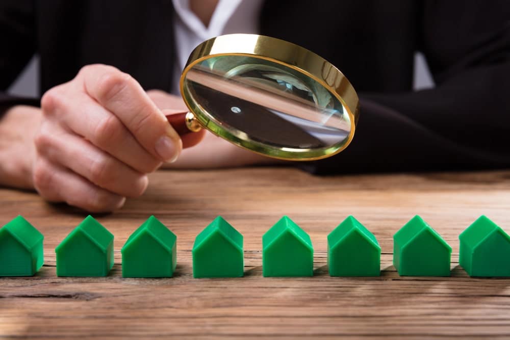 Image shows a person using a magnifying glass to inspect small green house models, concept of how an asset search reviews property ownership and financial records.