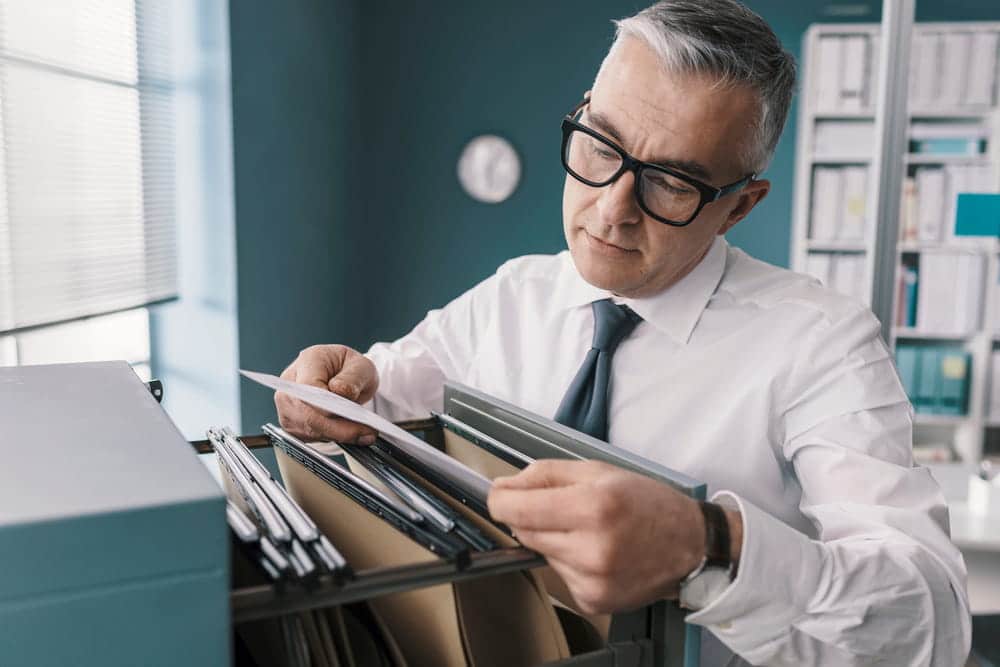 Image is of a professional reviewing physical file folders in an office setting, representing how documented records are examined during asset investigations