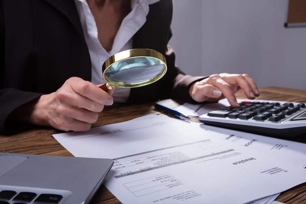 Image is of a professional reviewing financial paperwork with a magnifying glass, representing asset searches during detailed investigation.