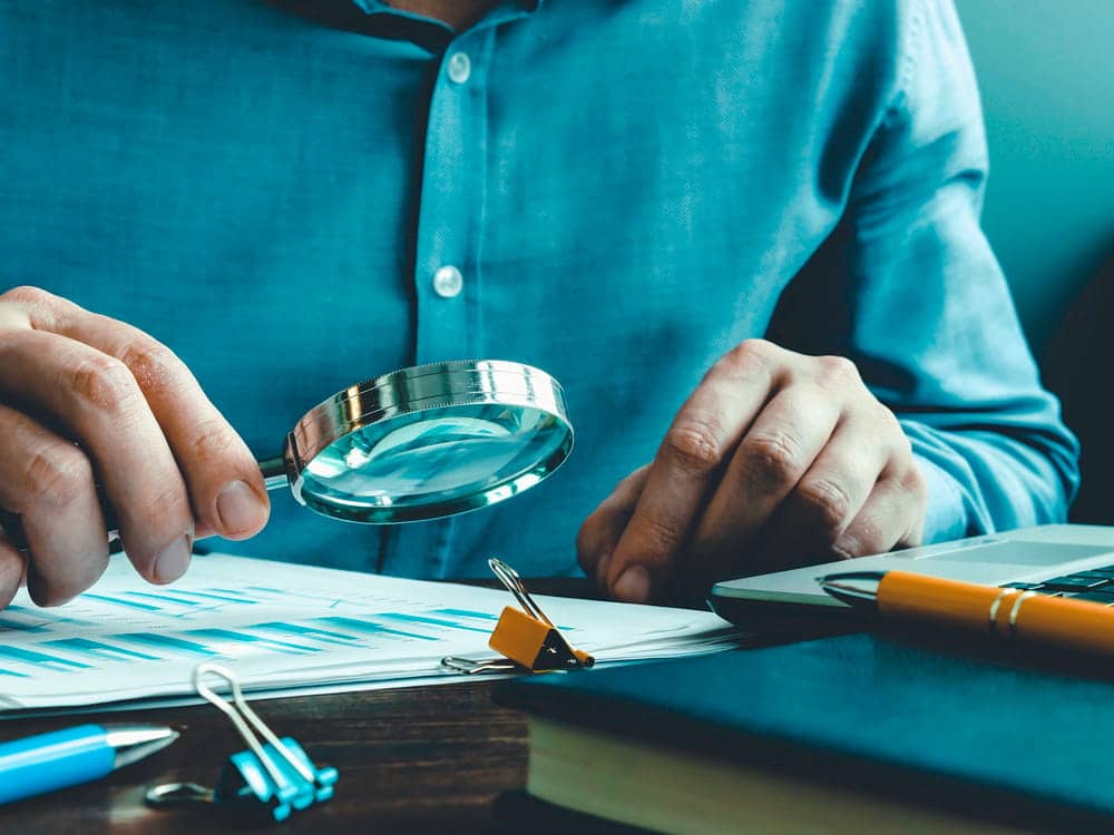 Image is of an investigator using a magnifying glass to examine documents, representing asset searches in uncovering corporate fraud