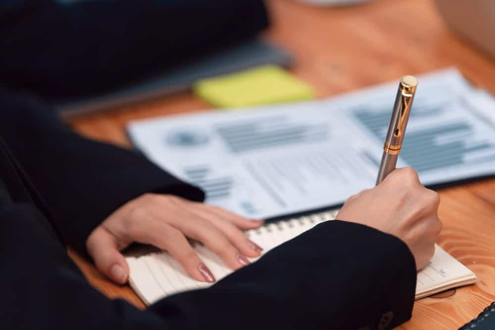 Image is of a person writing detailed notes beside financial documents, concept of compiling findings for an asset search report.