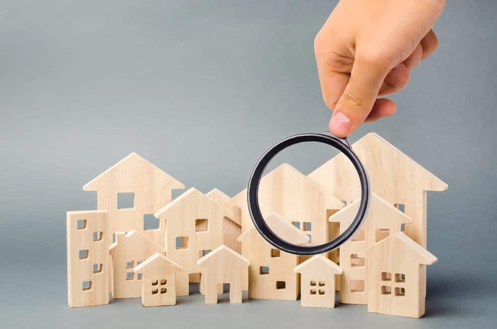 Image is of a magnifying glass examining miniature houses, concept of asset search identifying hidden real estate assets and ownership links.