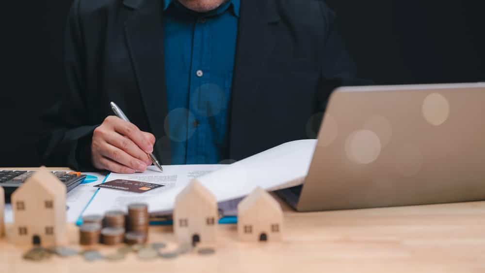 Image is of a man working on a laptop with documents and house models, concept of asset search tracing property records and financial connections.