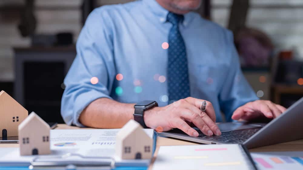 Image is of a professional reviewing financial documents with small house models, concept of asset search uncovering hidden real estate ownership.