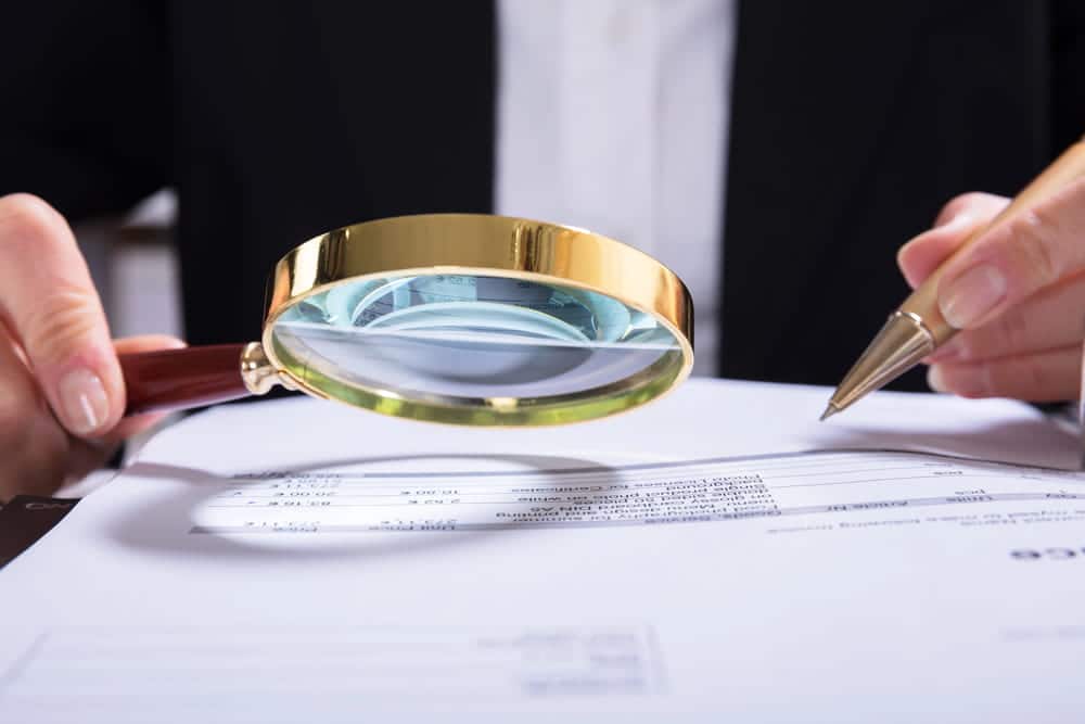 Image is of a person reviewing financial documents with a magnifying glass, representing the accuracy of asset search results in investigations